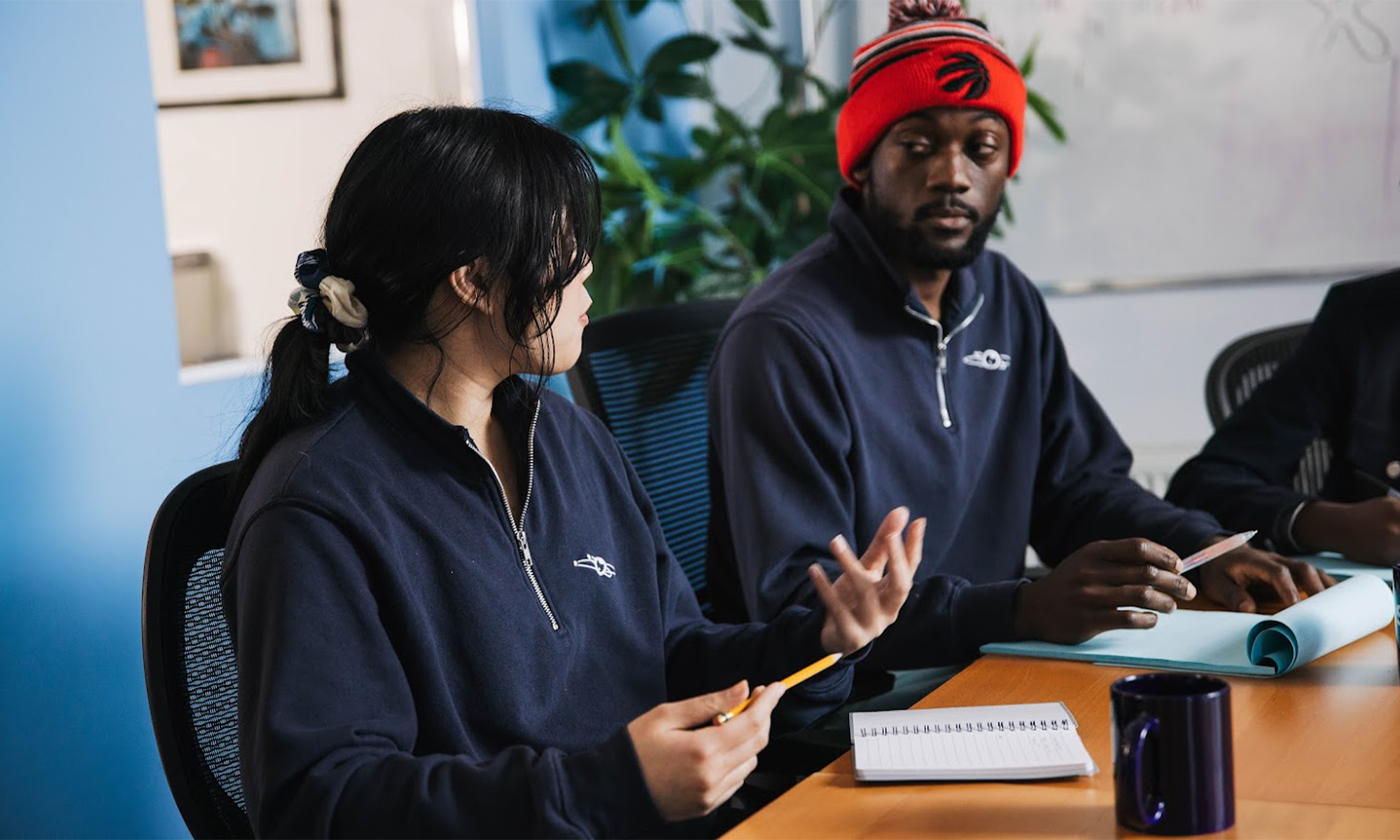 Two people in navy sweaters discuss at a table with notebooks, coffee mug, and papers in an office setting.