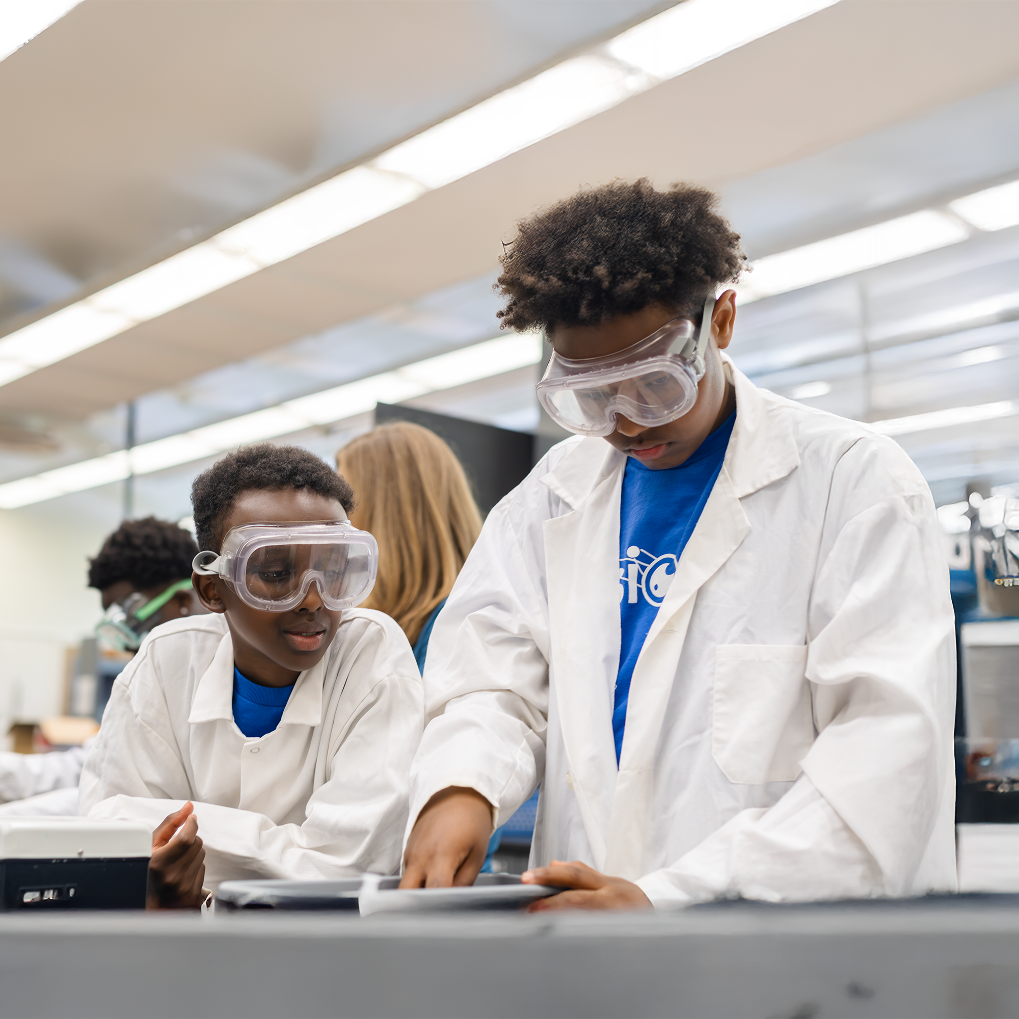 Two kids in lab coats and safety goggles working together on a science experiment in a laboratory classroom.
