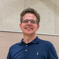 A smiling man with glasses wearing a navy blue polo shirt stands indoors against a beige wall.