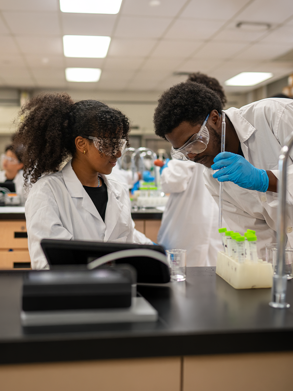 Two students in lab coats and goggles work with test tubes in a science laboratory.