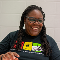Smiling woman with glasses and braids wearing a black T-shirt, sitting in front of a white brick wall.